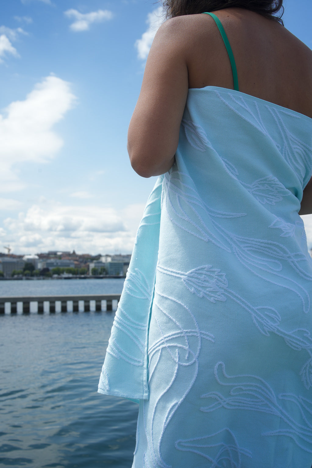 Person wrapped in a light aquasky towel with a white aquatic plant pattern, standing by a waterfront.