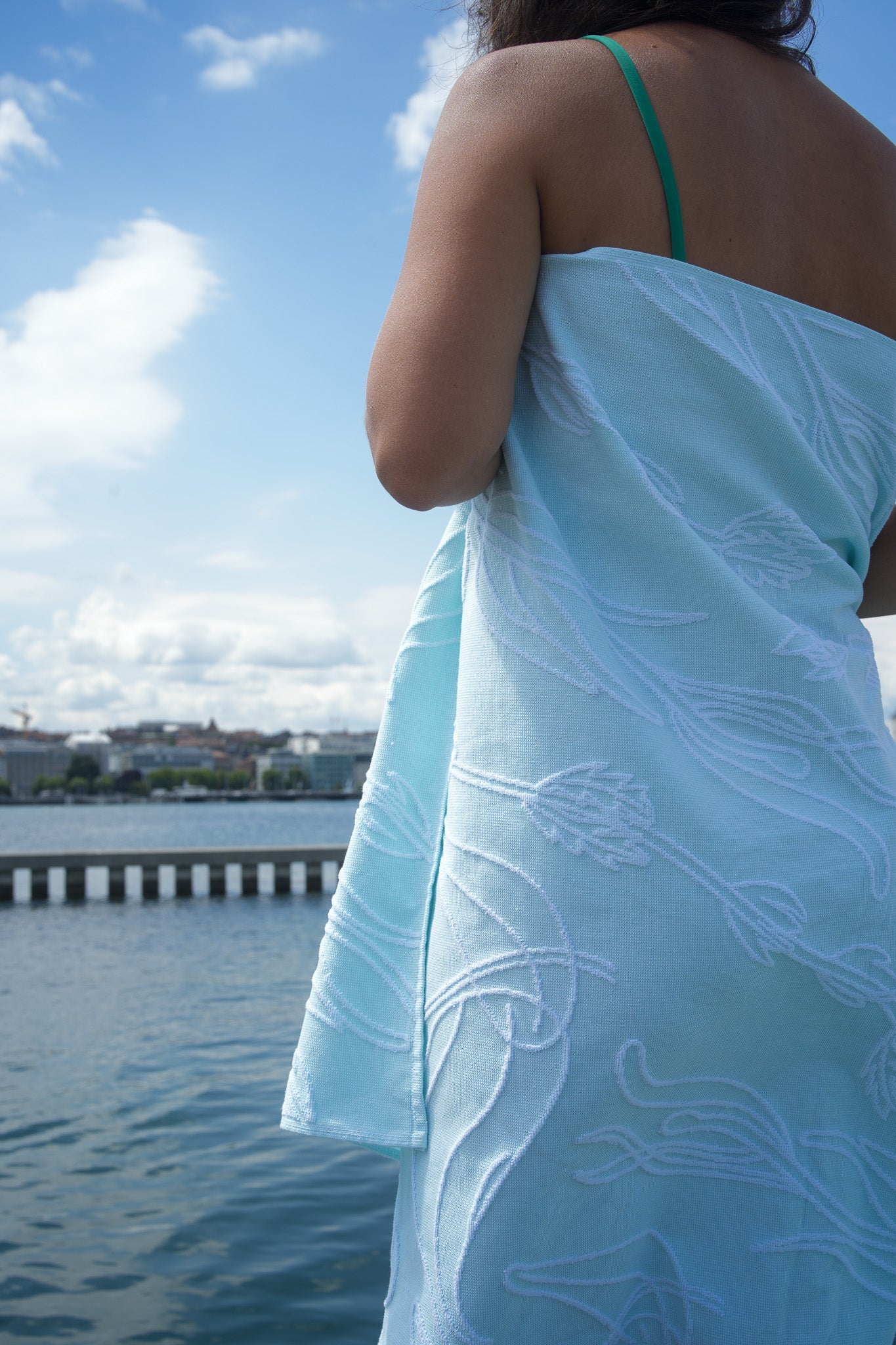 Person wrapped in a light aquasky towel with a white aquatic plant pattern, standing by a waterfront.
