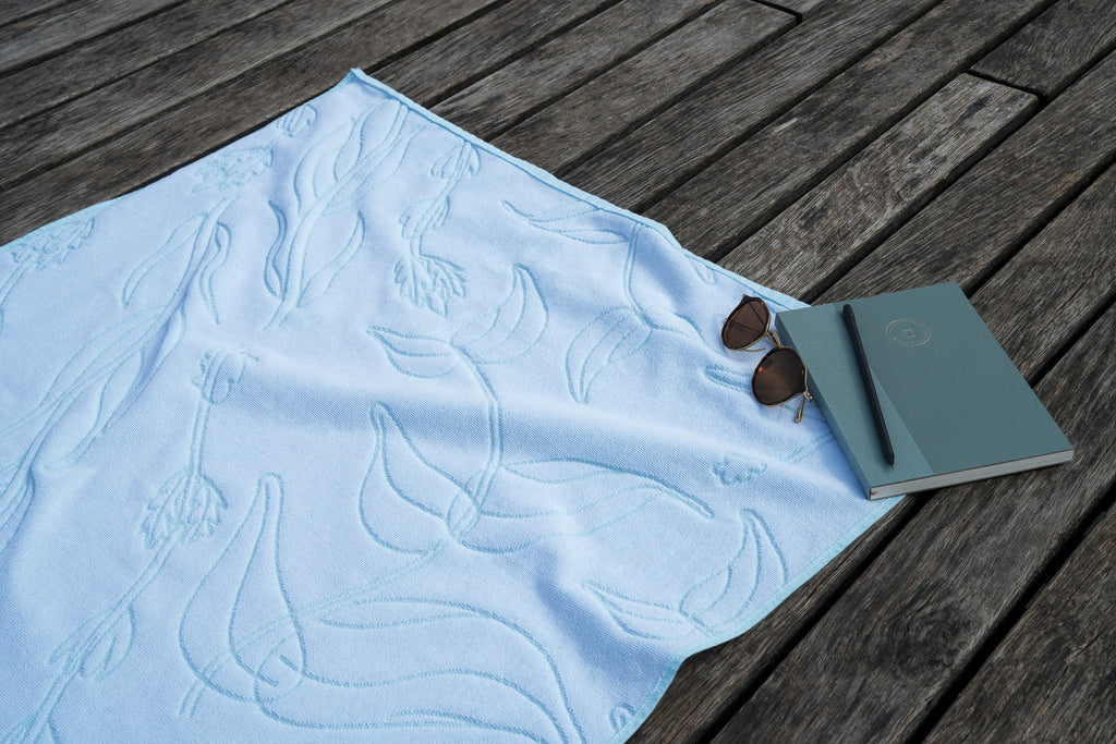 White textured beach towel with aquasky aquatic plant motif on a wooden deck, with a notebook, pencil and sunglasses.