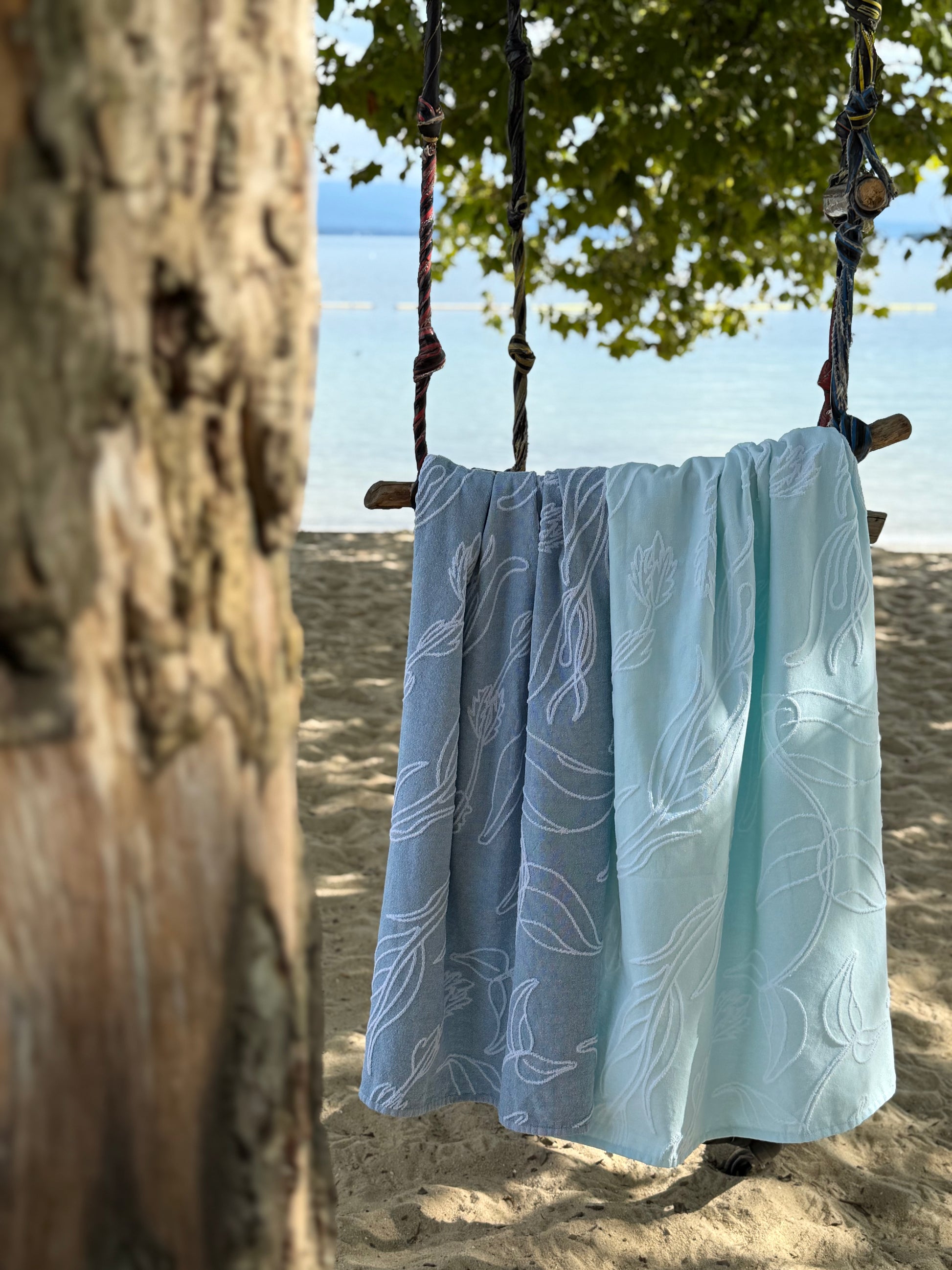 Light blue and light aquasky Vacationer towels with aquatic plant patterns hanging from a swing on the beach