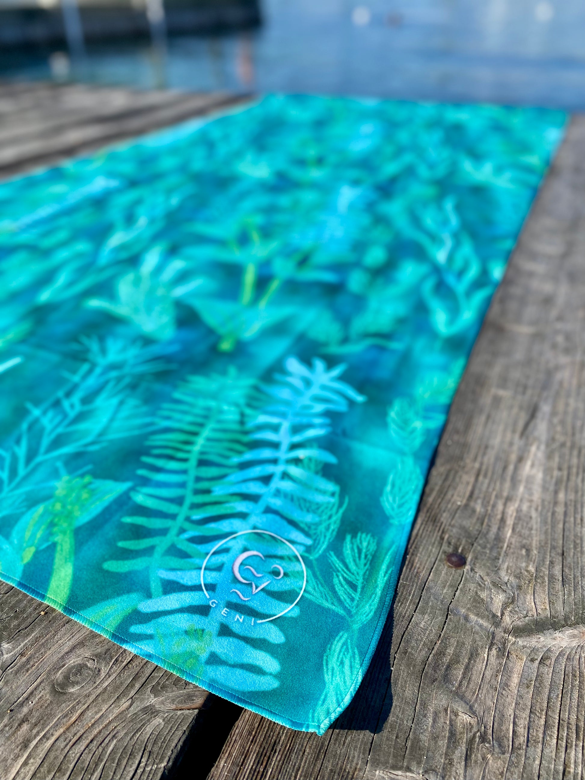 Blue-green Traveler beach towel with underwater plant pattern on a wooden deck by lake Geneva. Geni logo in the bottom right corner of the towel is visible in the foreground.