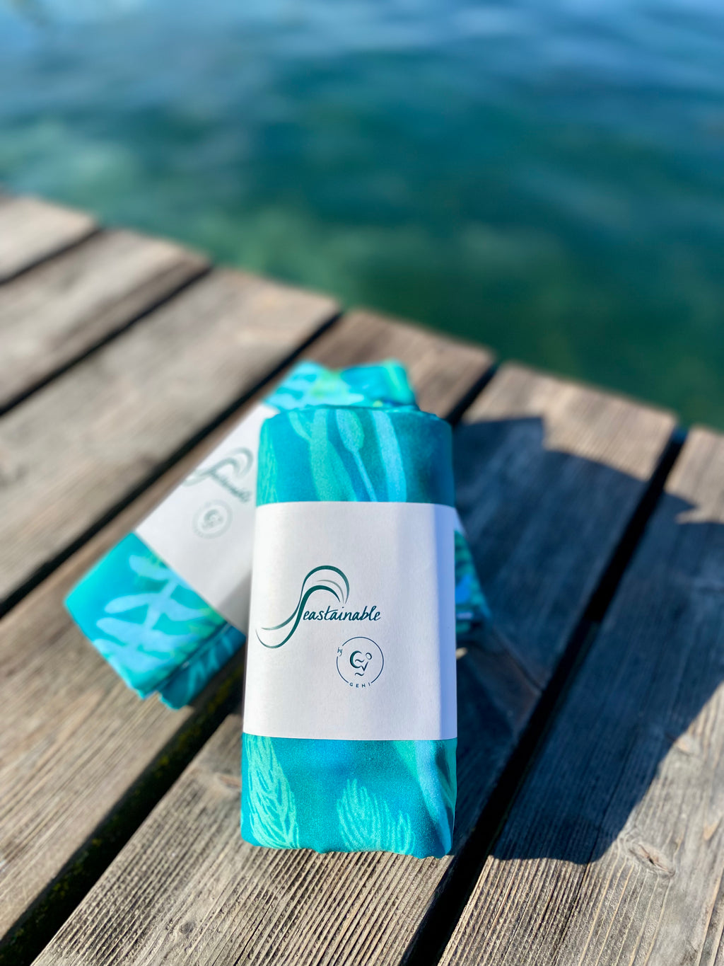 Blue-green Traveler beach towels with underwater plant pattern are wrapped with a paper packaging tag that says "Seastainable by Geni" on a wooden deck by lake Geneva.