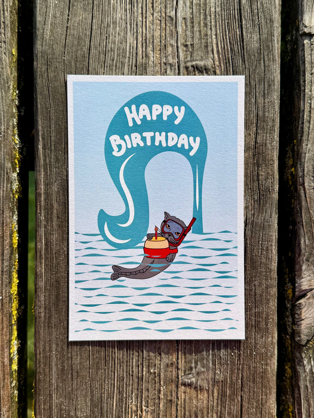 Illustration of catfish Darsea whofloats in the water holding a fondue pot with a candle in front of the Jet d'Eau of Geneva with the writing 'Happy Birthday' 