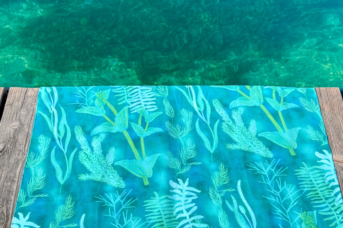 Blue-green Traveler beach towel with underwater plant pattern on a wooden deck by lake Geneva. Lake and aquatic plants in the background.