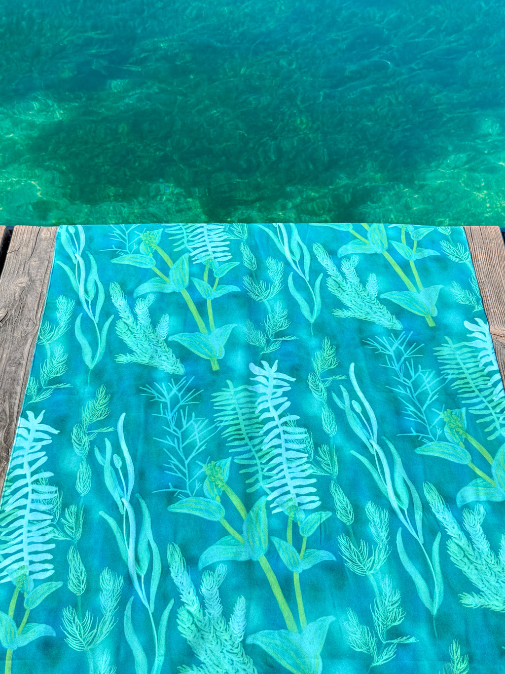Blue-green Traveler beach towel with underwater plant pattern on a wooden deck by lake Geneva. 