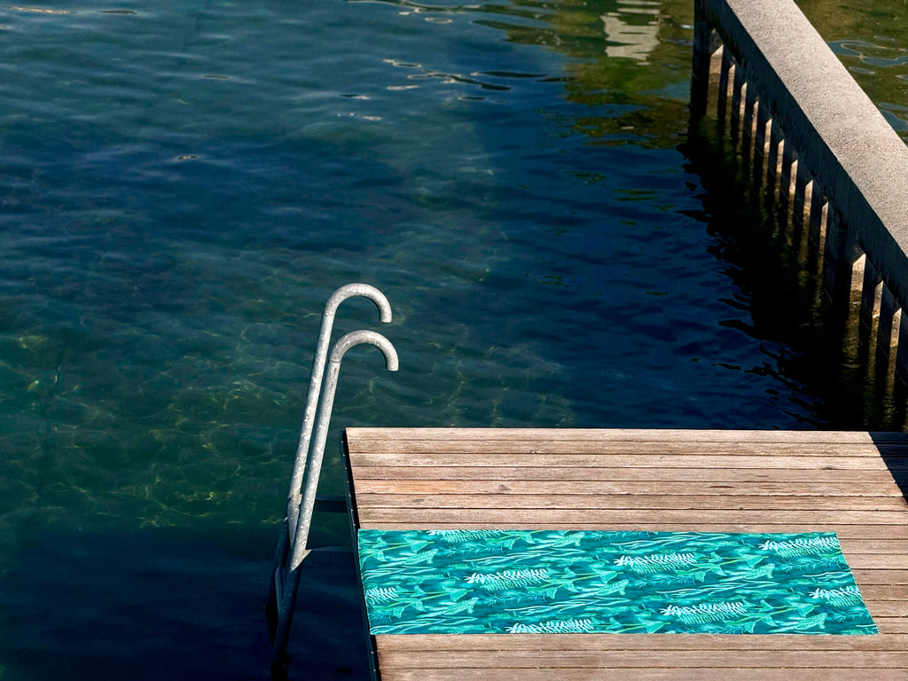 Blue-green Traveler beach towel with underwater plant pattern on a wooden deck by lake Geneva. 