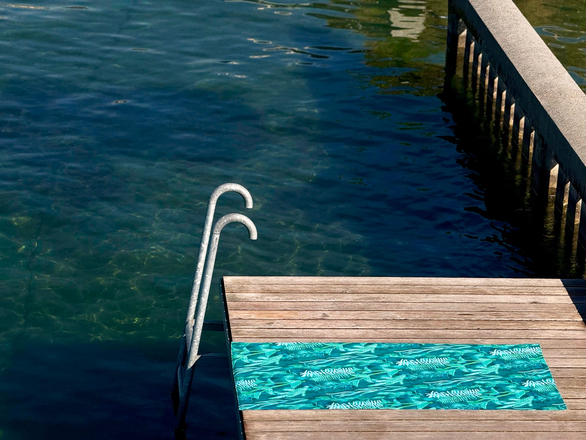 Blue-green Traveler beach towel with underwater plant pattern on a wooden deck by lake Geneva. 