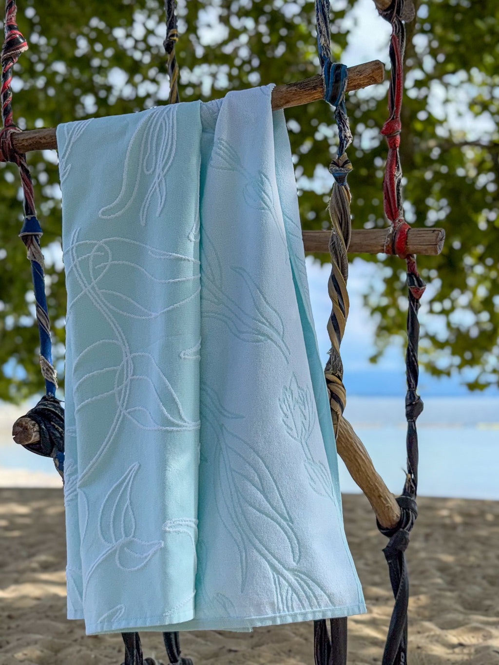 Light aquasky towel with white aquatic plant patterns hanging on a wooden swing against a water background.