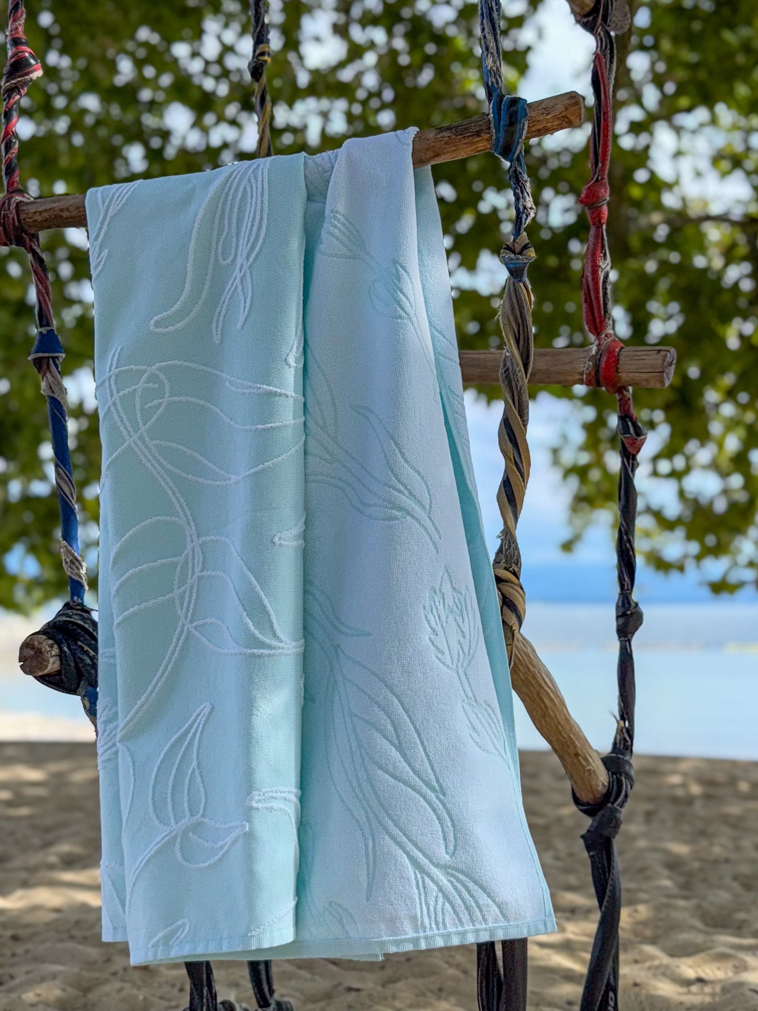 Light aquasky towel with white aquatic plant patterns hanging on a wooden swing against a water background.