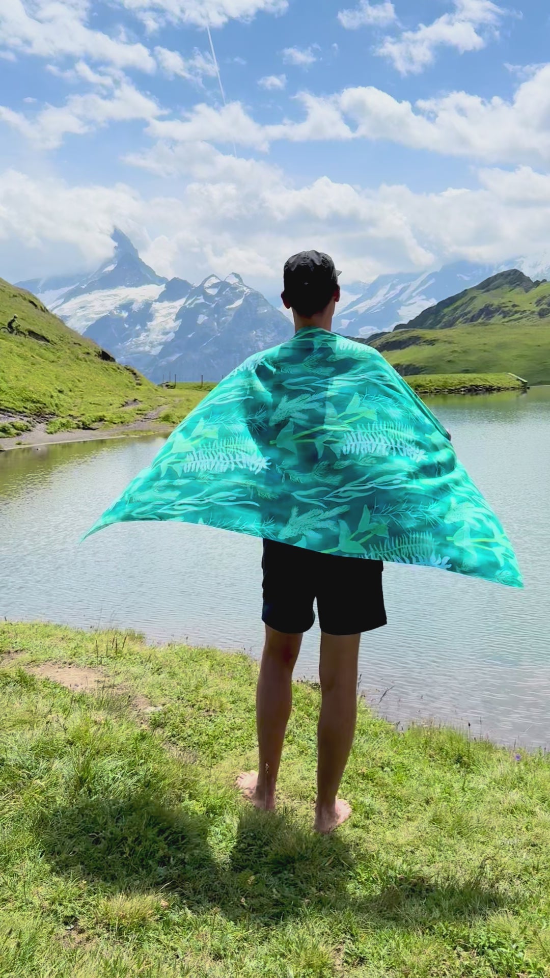 Blue-green Traveler beach towel with underwater plant pattern is wrapped around a man's shoulders and fluttering in the wind, against a mountain and lake scenic background.